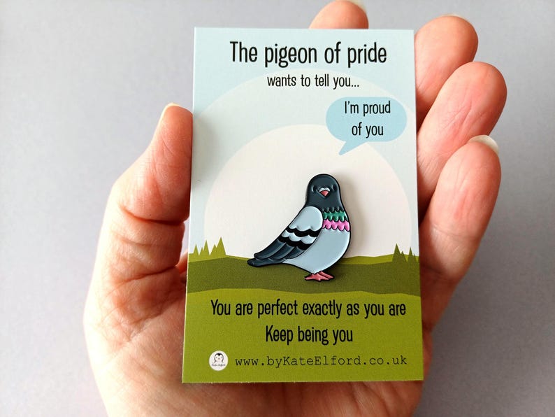 A pin badge depicting a cartoon pigeon with a light blue body, black wings, and pink and green details. The badge is on a card with the text "The pigeon of pride wants to tell you... I'm proud of you." and "You are perfect exactly as you are. Keep being you."