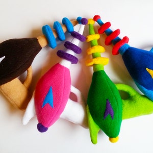 May include: Four plush toy phaser guns in different colors: brown, pink, green, and blue. Each phaser has a white star trek logo and a spiral detail on the back.