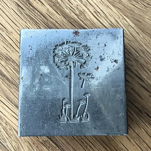 Könnte beinhalten: Ein quadratischer Metallstempel mit einem geprägten Design einer Löwenzahnblume mit langem Stiel, kleinen Grashalmen an der Basis und einem einzelnen schwebenden Samen. Die Oberfläche des silberfarbenen Metalls zeigt Gebrauchsspuren.
