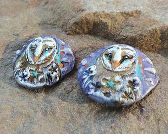 May include: Two round ceramic earrings with a purple background and a white owl design. The owls have orange and blue accents and a white star.