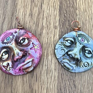 May include: Two circular clay pendants featuring sculpted faces with celestial motifs. The left pendant is painted in shades of pink and purple with star accents. The right pendant is painted in metallic blue and grey. Both have copper wire loops.