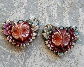 May include: Two heart-shaped owl pendants. Each pendant features a detailed owl face with large eyes and a textured surface. The owls are primarily red and orange, with accents of purple, blue, and white. They have a metal loop at the top for hanging.