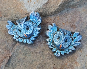 May include: Two blue owl-shaped decorative objects with detailed features. The owls have large, round eyes and are painted in shades of blue with orange accents. The objects are placed on a textured, light brown surface.