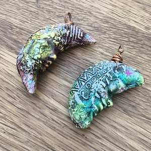 May include: Two crescent moon pendants featuring textured surfaces with iridescent green, blue, and purple hues. Each pendant has a copper wire loop at the top and displays intricate geometric patterns etched into the clay-like material.