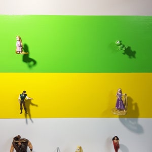 Könnte beinhalten: Eine Sammlung von Disney-Prinzessinnen- und Figuren, darunter Rapunzel, Flynn Rider, ein grünes Chamäleon, ein Pferd, ein Troll und eine brünette Frau in einem roten Kleid. Die Figuren sind auf einer weißen Oberfläche mit grünem, gelbem und weißem Hintergrund angeordnet.