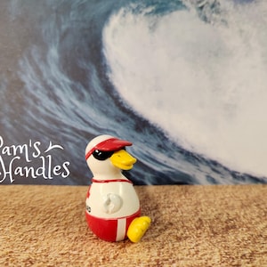 Pode incluir: Uma pequena figura de pato, usando óculos de sol, viseira vermelha e uniforme de salva-vidas. O pato é branco com detalhes em vermelho e pés amarelos. O fundo apresenta uma grande onda do oceano e o texto "Pam's Handles".