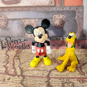 May include: Two Disney figurines: Mickey Mouse and Pluto. Mickey Mouse is in a classic pose, wearing red shorts, yellow shoes, and white gloves. Pluto is a yellow dog with a black ear and tongue sticking out. The figures are on a map.