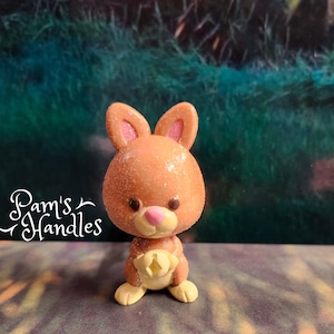 Puede incluir: Una pequeña figurita de conejo marrón brillante con orejas rosas y vientre amarillo. El conejo está de pie sobre una superficie marrón claro.