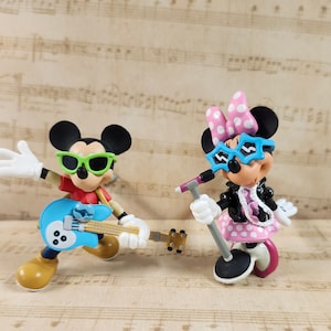 Op de afbeelding: Een blauwe en groene gitaar met een microfoonstandaard en twee cartoonfiguren. De figuren zijn Mickey Mouse en Minnie Mouse, die een zonnebril dragen en muziekinstrumenten vasthouden.