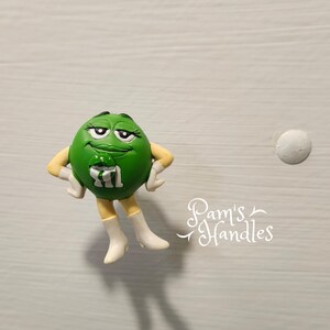 Peut inclure: Figurine verte repr&eacute;sentant un personnage M&M's, les bras crois&eacute;s, portant des bottes et des gants blancs. Le bonbon porte un "M" blanc sur le devant. Il s'agit d'un objet de fantaisie, peut-&ecirc;tre une poign&eacute;e de meuble.