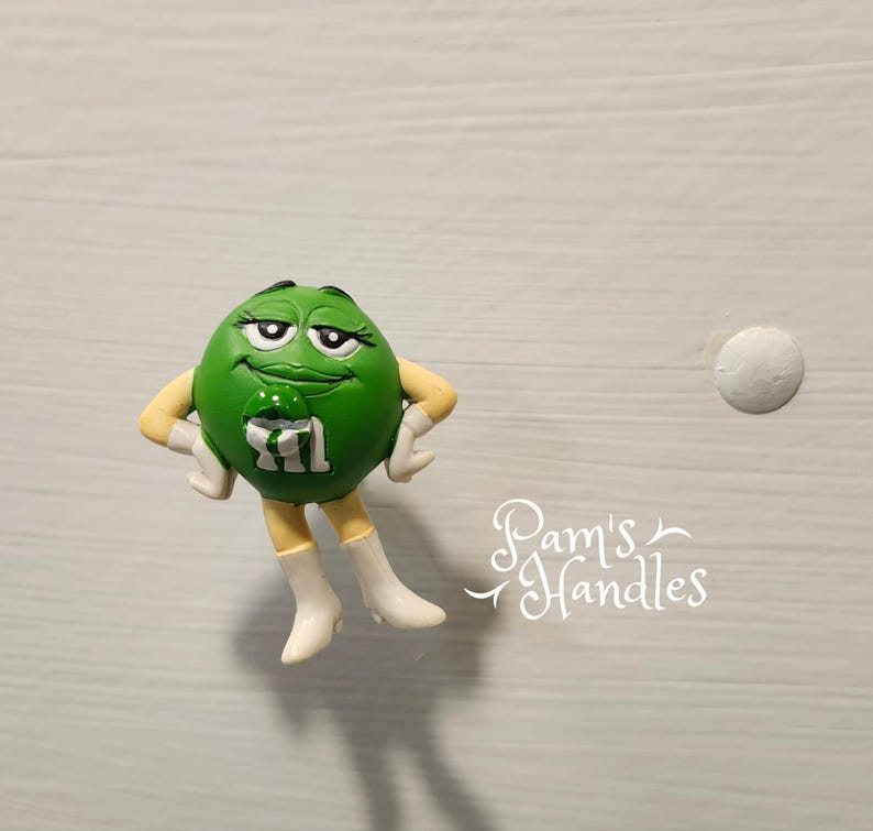 Peut inclure: Figurine verte repr&eacute;sentant un personnage M&M's, les bras crois&eacute;s, portant des bottes et des gants blancs. Le bonbon porte un "M" blanc sur le devant. Il s'agit d'un objet de fantaisie, peut-&ecirc;tre une poign&eacute;e de meuble.