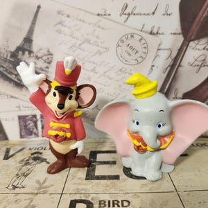 Può includere: Due statuette Disney: un topo in uniforme rossa e un elefante grigio con orecchie rosa. Il topo saluta e l'elefante ha un cappello giallo. Entrambi sono su una superficie a motivi.