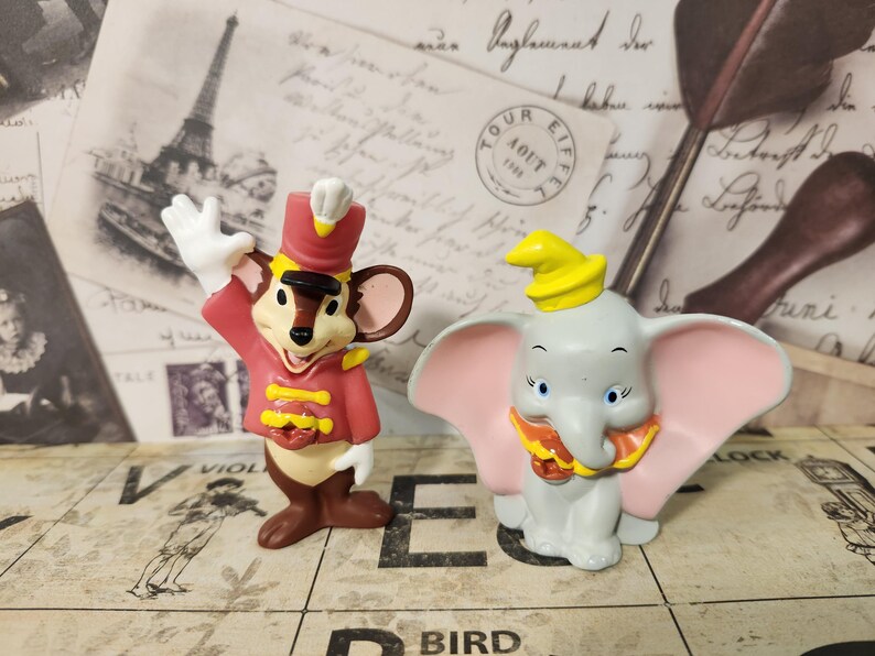 K&ouml;nnte beinhalten: Zwei Disney-Figuren: eine Maus in roter Uniform und ein grauer Elefant mit rosa Ohren. Die Maus winkt, und der Elefant hat einen gelben Hut. Beide stehen auf einer gemusterten Oberfl&auml;che.