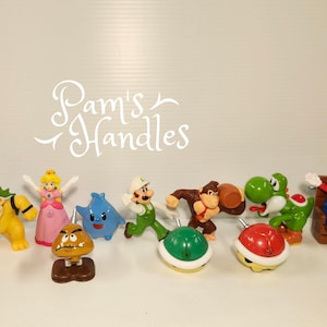 May include: A collection of ten colorful character-themed drawer knobs featuring figures from the Super Mario franchise, including Mario, Peach, Bowser, Donkey Kong, Yoshi, a Goomba, a green shell, a red shell, and a blue Luma, with the text Pam's Handles.
