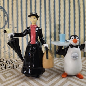 Peut inclure: Une figurine noire et blanche de Mary Poppins tenant un parapluie noir et un sac brun. Une figurine de pingouin en smoking tient un plateau avec un verre bleu.