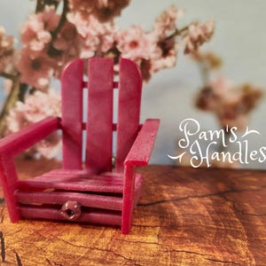 Pode incluir: Uma pequena cadeira Adirondack magenta com encosto de ripas e apoios de braços. A cadeira está colocada sobre um fundo desfocado de flores cor-de-rosa e uma superfície de madeira. O texto "Pam's Handles" é visível.