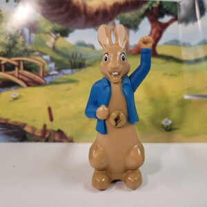 Könnte beinhalten: Eine braune Hasenfigur, die eine blaue Jacke trägt und den rechten Arm erhoben hat. Der Hase hat einen weißen Bauch und einen fröhlichen Gesichtsausdruck.