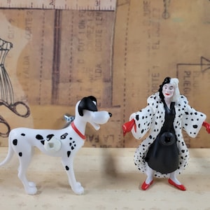 Könnte beinhalten: Eine schwarz-weiße Dalmatiner-Hundefigur und eine Cruella de Vil-Figur. Cruella trägt ein schwarzes Kleid und einen weiß-schwarz gefleckten Pelzmantel. Sie hat rote Schuhe und rote Handschuhe. Der Dalmatiner steht auf allen vieren mit eingerolltem Schwanz.