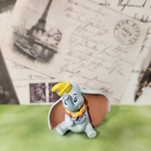 May include: A Dumbo the elephant figurine, seated. The figure is light blue with large, tan ears, a yellow hat, and a red and yellow collar. The background features vintage postcards and a pen.