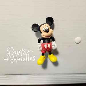 以下が含まれることがあります： 黒い耳、黒い胴体、赤いショートパンツ、白い手袋、黄色の靴を履いたミッキーマウスのフィギュア。フィギュアは明るい灰色の表面に立っています。「Pam's Handles」というテキストは白い筆記体です。