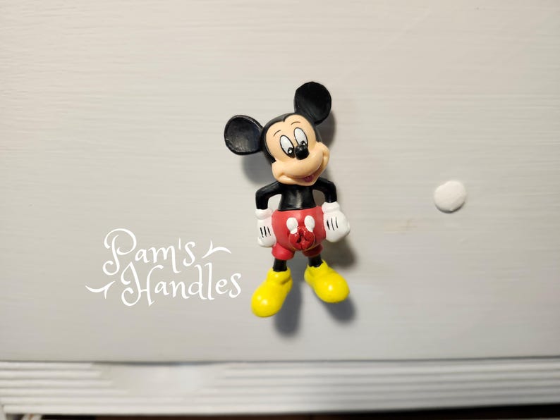 以下が含まれることがあります： 黒い耳、黒い胴体、赤いショートパンツ、白い手袋、黄色の靴を履いたミッキーマウスのフィギュア。フィギュアは明るい灰色の表面に立っています。「Pam's Handles」というテキストは白い筆記体です。