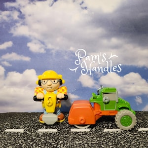 May include: A yellow toy construction worker figure with a yellow scooter and a green and orange road roller. The background is a blue sky with clouds. The text "Pam's Handles" is in the upper right corner.