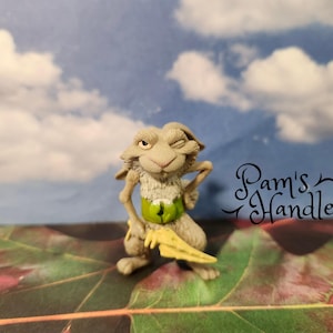 Könnte beinhalten: Eine skurrile Figur eines zwinkernden, kaninchenartigen Wesens mit grünem Bauch und gelbem Gegenstand. Die Figur ist vor dem Hintergrund eines bewölkten Himmels und Herbstblättern platziert. Der Text "Pam's Handles" ist sichtbar.