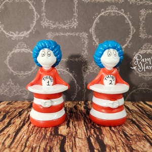 Peut inclure: Deux figurines de Chose 1 et Chose 2 du livre "Le Chat chapeauté" du Dr. Seuss. Chaque figurine a une tête bleue et duveteuse, un visage blanc et une chemise orange avec un cercle blanc. La base est rayée rouge et blanche.
