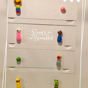 Puede incluir: Cómoda blanca con seis cajones con tiradores en forma de animales de colores. Los tiradores incluyen un león, un oso, un perro, una cebra, un gato y una oruga. La cómoda está etiquetada como "Pam's Handles".