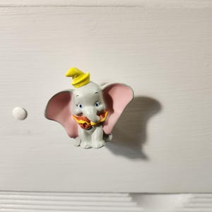 K&ouml;nnte beinhalten: Ein Dumbo-Schrankknopf. Der Knopf zeigt einen grauen Elefanten mit gro&szlig;en rosa Ohren, einem gelben Hut und einem rot-gelben Kragen. Der Knopf ist auf einer wei&szlig;en Oberfl&auml;che montiert.