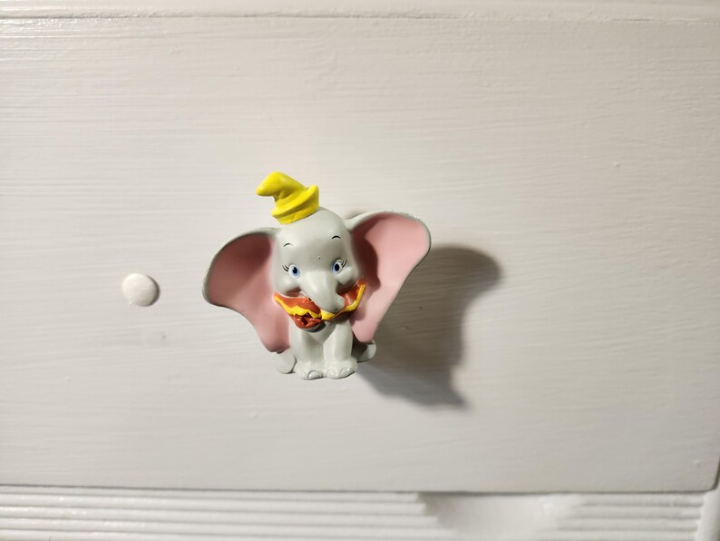 K&ouml;nnte beinhalten: Ein Dumbo-Schrankknopf. Der Knopf zeigt einen grauen Elefanten mit gro&szlig;en rosa Ohren, einem gelben Hut und einem rot-gelben Kragen. Der Knopf ist auf einer wei&szlig;en Oberfl&auml;che montiert.