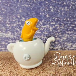 May include: A novelty white teapot with a yellow fish figurine popping out. The fish has a surprised expression. The teapot has a small handle and spout. The background is a textured blue and white pattern. The text "Pam's Handles" is in the lower right corner.