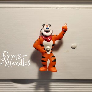 以下が含まれることがあります： 赤いバンダナをつけたオレンジと黒の虎の漫画キャラクターが指を立てています。バンダナには「トニー」と書かれています。キャラクターはプラスチック製のフィギュアです。