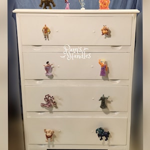 Può includere: Una cassettiera bianca con cinque cassetti. Ogni maniglia del cassetto è una diversa statuetta di un personaggio Disney. Il cassetto superiore ha una statuetta dorata, una statuetta viola, una statuetta blu e una statuetta arancione. Il secondo cassetto ha una statuetta di un uomo in armatura, una statuetta di un animale dei cartoni animati e il testo "Pam's Handles". Il terzo cassetto ha una statuetta viola, una statuetta di un uomo con una tunica e il testo "Pam's Handles". Il quarto cassetto ha una statuetta rosa di un drago e una statuetta di un uomo con una tunica. Il quinto cassetto ha una statuetta di un uomo con un pannolino e una statuetta di un uomo che cavalca un cavallo.