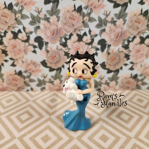 May include: A blue and white ceramic figurine of a cartoon character holding a white puppy. The character has black hair and a blue dress. The figurine is on a beige and white tiled surface.