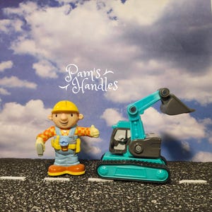 May include: A toy construction worker figure with a yellow hard hat, orange and yellow plaid shirt, and blue overalls gives a thumbs up next to a teal and gray toy excavator. The text "Pam's Handles" is in the background.