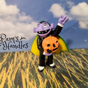 May include: A figurine of a cartoon vampire holding an orange jack-o'-lantern. The vampire has purple skin, a black suit, and a yellow and green cape. The figurine is set against a backdrop of a blue sky with clouds and tall grass. The text "Pam's Handles" is in the image.