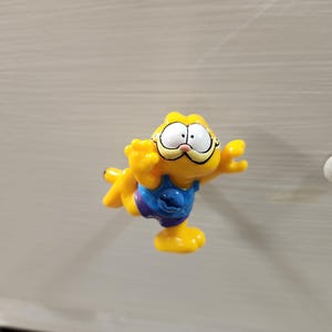 May include: A small yellow Garfield figurine depicted in a dynamic pose with arms outstretched. The character wears a blue tank top and purple shorts, featuring large white eyes, a pink nose, and a wide grin, captured in a bright, cartoonish style.