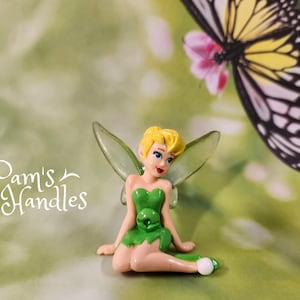 Peut inclure: Figurine d'une fée aux cheveux blonds, vêtue d'une robe verte et d'ailes translucides. La fée est assise sur une surface verte et blanche. Un grand papillon aux ailes jaunes et noires est en arrière-plan. Le texte "Pam's Handles" est dans l'image.