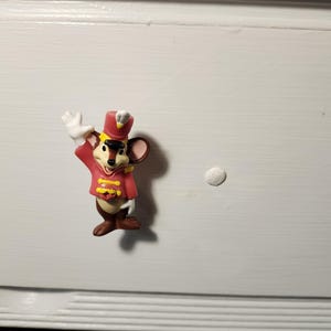 K&ouml;nnte beinhalten: Kleine Figur einer Maus in roter und gelber Uniform, die mit einer wei&szlig; behandschuhten Hand winkt. Die Maus hat braunes Fell, einen roten Hut und steht auf einer wei&szlig;en Oberfl&auml;che. Die Figur ist ein dekoratives Objekt.