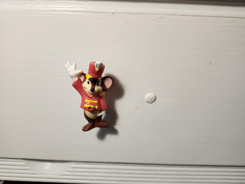 K&ouml;nnte beinhalten: Kleine Figur einer Maus in roter und gelber Uniform, die mit einer wei&szlig; behandschuhten Hand winkt. Die Maus hat braunes Fell, einen roten Hut und steht auf einer wei&szlig;en Oberfl&auml;che. Die Figur ist ein dekoratives Objekt.