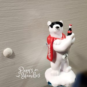 Peut inclure: Poignée de meuble représentant un ours polaire blanc portant des lunettes de soleil et une écharpe rouge Coca-Cola, tenant une petite bouteille de Coca-Cola. La poignée est montée sur une base blanche.
