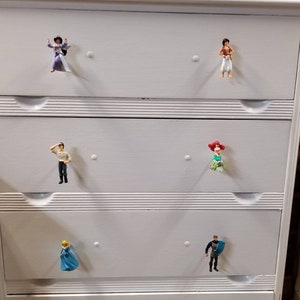May include: White chest of drawers with four drawers. Each drawer pull is a different plastic figurine. The top drawer has a purple figurine, the second drawer has a brown figurine, the third drawer has a red figurine, and the bottom drawer has a blue figurine.