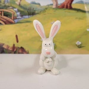 May include: A white ceramic bunny figurine with pink ears and a pink nose. The bunny is holding a white egg in its paws.