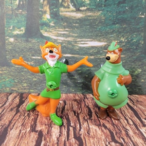 Könnte beinhalten: Zwei Plastikfiguren von Charakteren aus dem Disney-Film Robin Hood. Der Fuchs trägt eine grüne Tunika und einen braunen Hut. Der Bär trägt eine grüne Tunika und einen braunen Hut. Beide Figuren stehen auf einer Holzoberfläche.