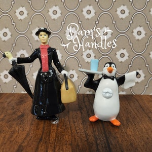 May include: A black and white figurine of Mary Poppins holding an umbrella and a yellow suitcase. A black and white penguin figurine is holding a tray with a blue glass. The figurines are on a brown wooden surface.