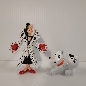 Könnte beinhalten: Eine Plastikfigur von Cruella de Vil aus dem Disney-Film 101 Dalmatiner. Sie trägt einen schwarz-weißen Dalmatiner-Mantel mit roten Handschuhen und roten Schuhen. Sie steht neben einem weißen Dalmatiner-Welpen mit schwarzen Flecken.