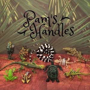 May include: A collection of green, brown and black animal figurines, including a snake, a turtle, a frog, a lizard and a crocodile. The figurines are arranged on a brown surface with a green leafy background. The text "Pam's Handles" is written in black cursive on the background.