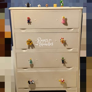 May include: A white four-drawer dresser featuring various Super Mario character figurines as drawer pulls. The second drawer from the top displays the white text Pam's Handles. Small character figures are also lined up on the top surface.