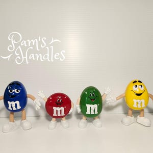 May include: Four colourful M&M's character figurines. The figurines are blue, red, green, and yellow, each with white gloves and shoes. The letter "m" is printed on the front of each character. The text "Pam's Handles" is in the background.